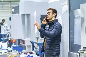 Communicative Young Adult Man Stampe: Engineer talking to the mobile in a computer numeric control factory