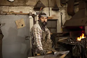 Blacksmith At Work Stampe: The portrait of bearded blacksmith preparing to work in his workshop